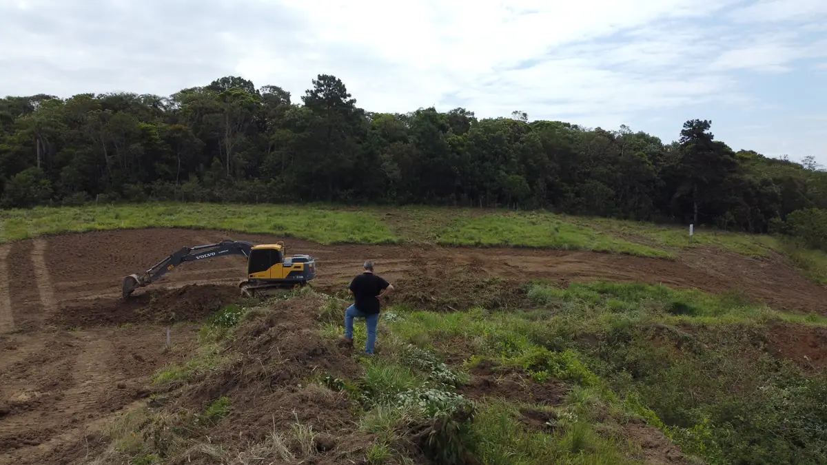 Escavadeira nivelando o terreno junto à mata, acompanhada por um profissional em campo